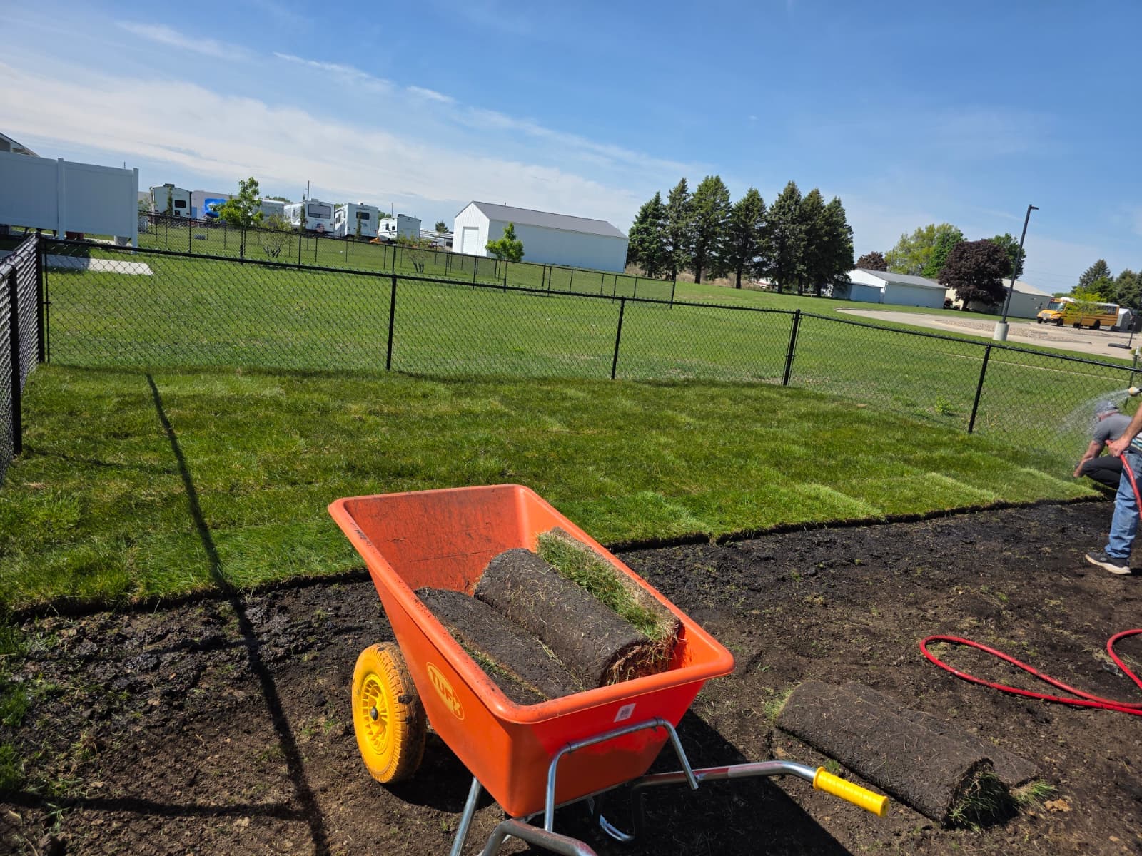 Rolled turf in a wheelbarrow beside an edged soil area and fenced lawn